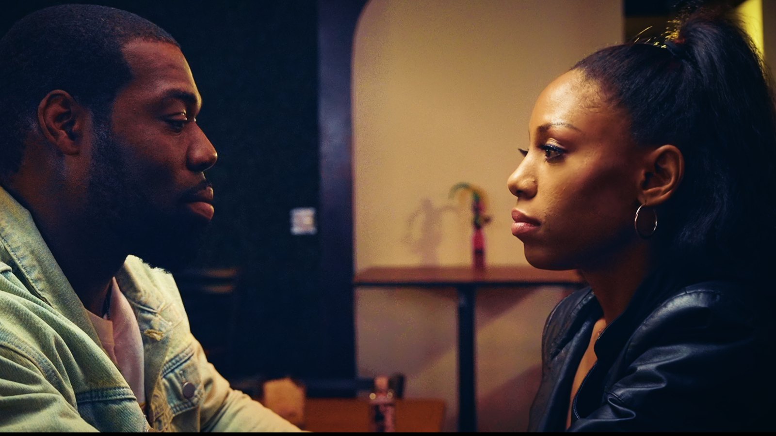 Reel Luv film still: two characters facing each other in profile in a dimly lit room, mid conversation